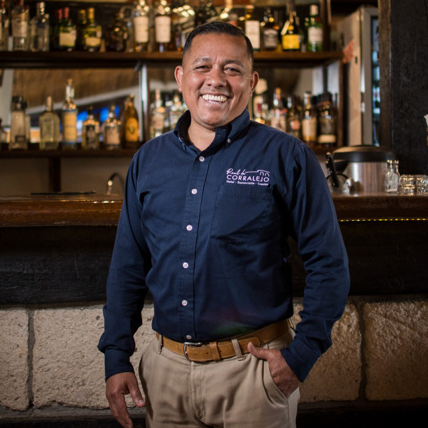 Hombre joven posando frente a una cantina con una camisa azul y pantalón café