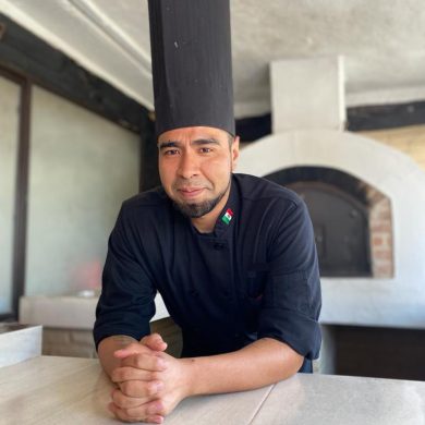 Chef joven frene a un horno de leña con filipina y gorra negra